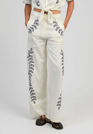 Pantalon large crème avec broderie de feuilles noires, mains dans les poches, associé à des sandales noires tissées et un haut court noué.