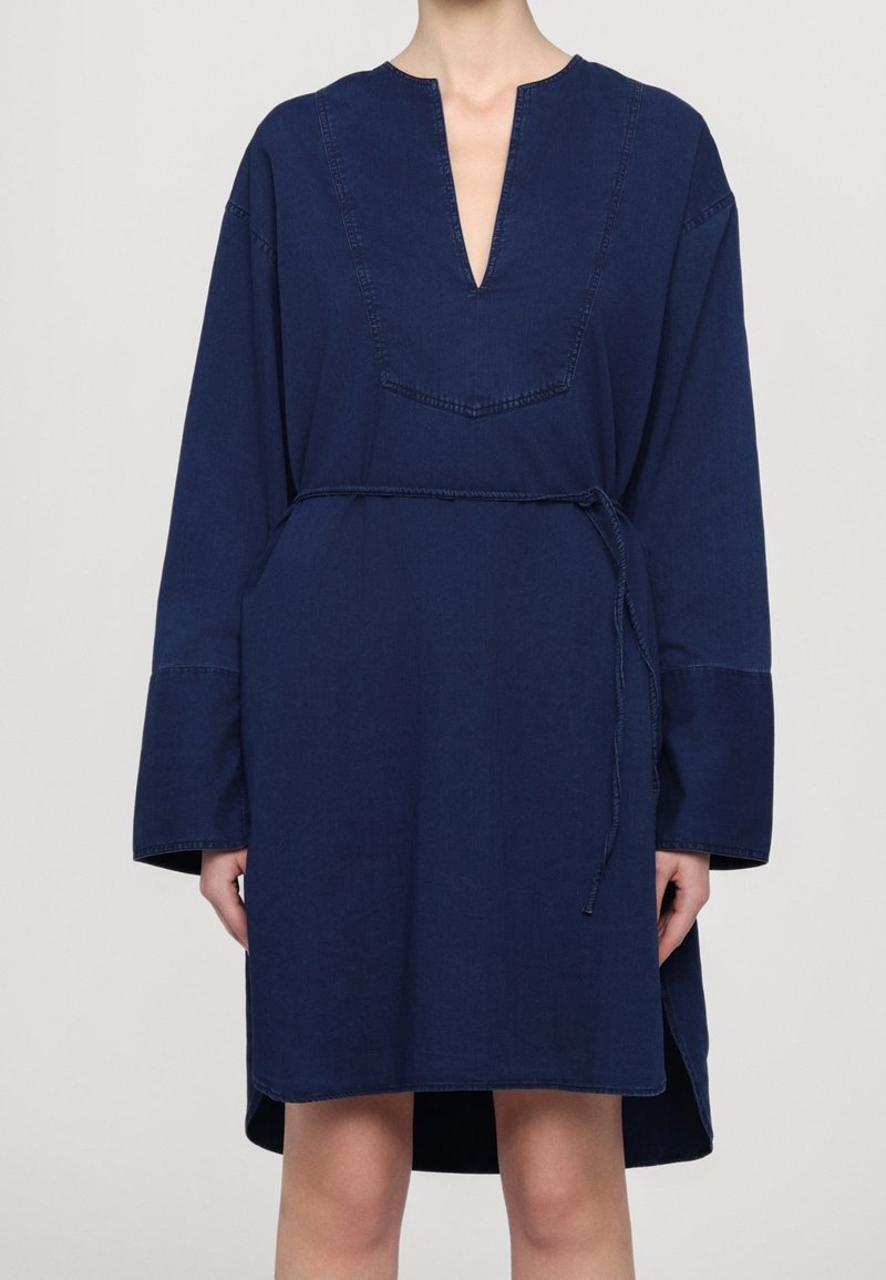 Kobieta ubrana w luźną granatową sukienkę z długimi rękawami, dekoltem w szpic, przeszyciami na panelu piersiowym oraz cienkim paskiem związanym w talii.