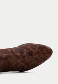 Botín de ante marrón hasta el tobillo con puntera afilada. Presenta un patrón de print de leopardo y una parte superior lisa y texturizada. Bordes curvados y tacón bajo.