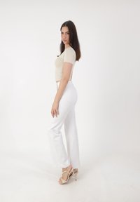 Mujer con cabello largo y oscuro, vestida con un top corto beige, pantalones blancos de cintura alta y sandalias de tacón alto beige, de pie contra un fondo blanco.