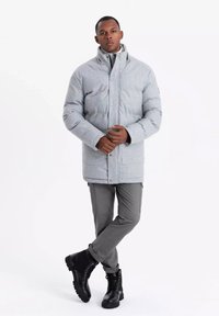 Abrigo acolchado gris con cuello alto, cierre de botones, llevado sobre pantalones grises. Botas negras con cordones y suela robusta. Fondo blanco liso.