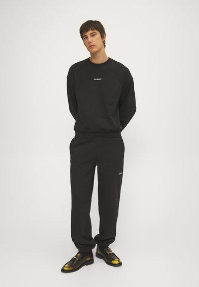 Melnas sporta džemperis un saskaņotas bikses ar brīvu piegriezumu. Satur vienkāršu tekstūru un diskrētu balto zīmolu. Spīguļojošas kurpes papildina kopskatu.