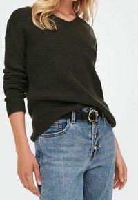 Pull en maille vert foncé avec un col en V, associé à un jean bleu taille haute. Ceinture noire avec une boucle ronde à motifs. Tissu texturé.