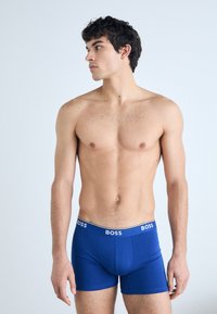 Blaue Boxer-Shorts aus dehnbarem Stoff mit einem weißen Logo-Bund. Das Kleidungsstück hat eine enganliegende Passform und eine glatte Textur.