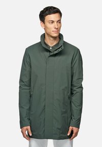 Giacca impermeabile verde scuro con colletto alto, tasche laterali e una texture liscia. Presenta una chiusura con zip e bottoni per un fissaggio sicuro.