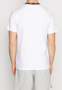 Vit t-shirt med rund halsringning och korta ärmar, med ett svart varumärkesband vid kragen och en mjuk bomullstextur.