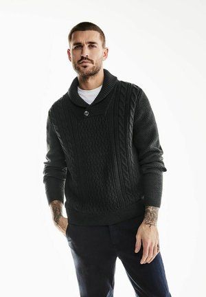 Pull noir en tricot avec un col châle, motif en torsades et détails de boutons. Le modèle le porte par-dessus une chemise blanche, associé à un pantalon foncé.