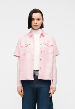 Kvinne med kort mørkt hår som har på seg en hvit turtleneck, rosa kortermet skjorte med knapper, mørkeblå jeans og et hjerteformet beltespenne.
