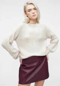 Blondynka stoi z rękami na biodrach, ubrana w beżowy sweter dziany i bordową skórzaną spódnicę na białym tle.