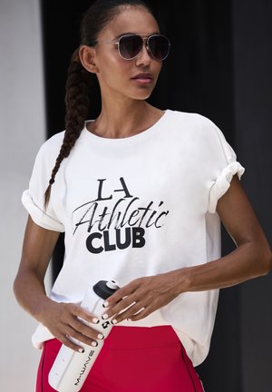 Woman with braided hair and sunglasses wearing "LA Athletic Club" white t-shirt and red leggings, holding a white water bottle.