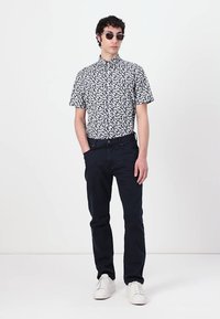 Camicetta a maniche corte con un motivo di foglie in bianco e nero, abbinata a jeans scuri e sneakers bianche, indossata con occhiali da sole rotondi.
