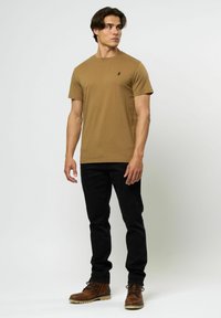 Un tricou scurt maro cu mâneci scurte, guler rotund și un mic logo negru pe piept, purtat cu blugi negri și cizme maro cu șireturi.
