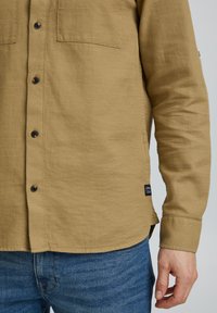 Chemise kaki à manches longues en tissu texturé, dotée de deux poches poitrine, de boutons noirs et d'une étiquette au bas. Portée avec un jean bleu.
