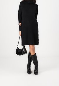 Robe noire en laine avec un col bénitier, associée à un petit sac à main noir en cuir verni et des bottes noires montantes à bouts pointus et à boucles.