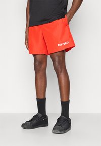 Κόκκινες αθλητικές βερμούδες με ελαφρύ σχεδιασμό, με λευκό λογότυπο και πλαϊνές τσέπες. Φορεμένες με μαύρες κάλτσες και αθλητικά παπούτσια.