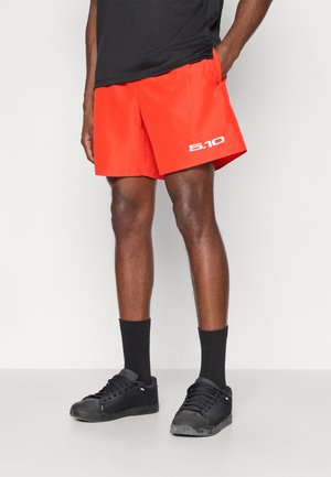 Calções atléticos vermelhos com um design leve, apresentando um logotipo branco e bolsos laterais. Usados com meias pretas e ténis.