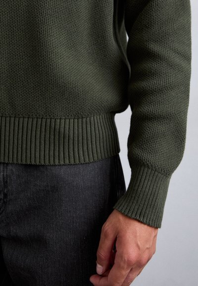 Strickpullover in Dunkelgrün mit strukturiertem Oberteil, gerippten Bündchen und Saum. Kombiniert mit dunkelgrauen Jeans, die filigrane Stickdetails zeigen.
