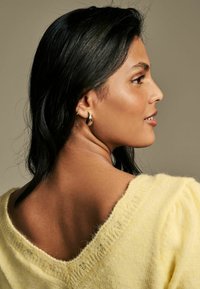 Maglione giallo lavorato a maglia con scollatura ampia, morbida texture e maniche a sbuffo. La modella ha i capelli lunghi neri e indossa orecchini d'oro.