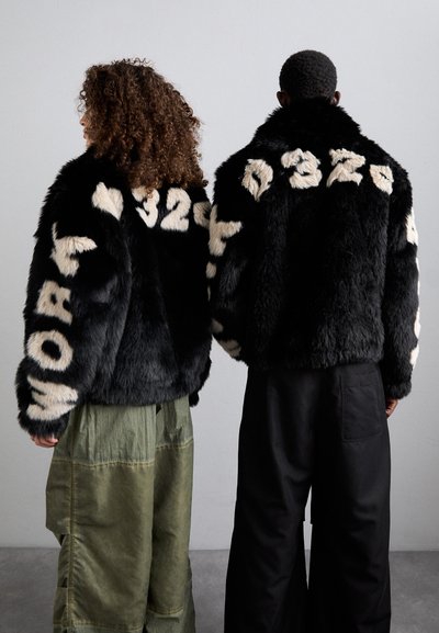 Dos personas que llevan chaquetas de pelaje sintético negro de gran tamaño con texto en crema en contraste. Las chaquetas presentan letras grandes en la espalda y en las mangas.