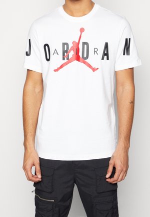 T-shirt blanc avec le texte « JORDAN » et le logo rouge du joueur de basket Jumpman centré sur la poitrine, porté avec un pantalon cargo noir.