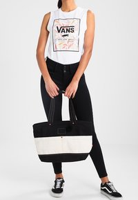 Bolso tote en negro y crema con asas dobles, que cuenta con costuras en contraste y un bolsillo en la parte frontal. El modelo lleva puesto una camiseta sin mangas gr�áfica blanca y jeans negros.