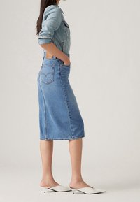 Falda midi de mezclilla en azul claro, con dobladillo sin terminar, dos bolsillos en la parte trasera y costuras en contraste. Combinada con zapatos blancos tipo slip-on.