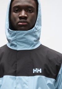 Φωτεινό μπλε και μαύρο μπουφάν με κουκούλα και ψηλό γιακά, με ρυθμιζόμενο κορδόνι στην κουκούλα και λογότυπο στο στήθος.