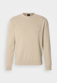 Beige színű, puha kötött anyagból készült pulóver, kerek nyakkivágással és bordás mandzsettákkal, valamint aljjal. Kis logó embléma a mellrészen.