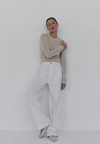 Beżowa bluzka z długim rękawem i dopasowanymi mankietami, zestawiona z szerokimi białymi spodniami i jasnymi czółenkami, na jednolitym tle.