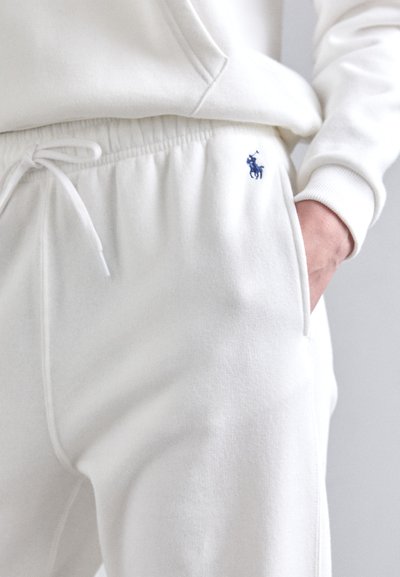 Vita bomullsbyxor med elastisk midja och dragsko, med en liten blå broderad logotyp på vänster sida.