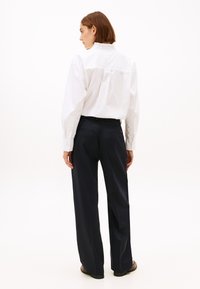 Wit overhemd met geplooide kraag en lange mouwen, gecombineerd met donkerblauwe broek met wijde pijpen. Glad stof met getailleerd ontwerp en achterzakken.