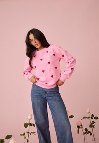 Žena s dlhými tmavými vlasmi, oblečená v ružovej mikine s červenými srdiečkami, modrých džínsach a veľkých slnečných okuliaroch, stojaca medzi ružovými ružami vo váze.