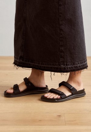 Fødder iført sorte sandaler med to justerbare stropper, parret med et langt sort denimsnørk med frynset kant, stående på et trægulv.