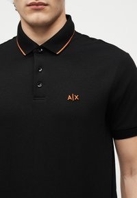 Homme portant un polo noir avec un logo A|X orange sur la poitrine et un bord orange assorti sur le col, sur un fond uni.