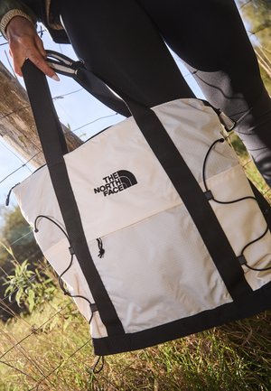 Saco de duffel de tecido bege com alças pretas, apresentando um logótipo, fecho de cordão e detalhes adicionais em corda na parte exterior.