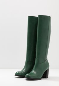 Bottes montantes en cuir vert jusqu'aux genoux avec un bout arrondi, une texture lisse et un talon épais. Dotées d'une fermeture éclair latérale et d'accents de couture.