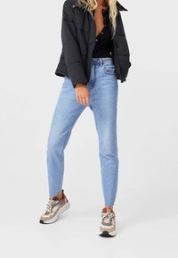 Zwarte pufferjack over een zwart top, blauwe skinny jeans en beige sneakers met accenten. Het materiaal lijkt soepel; het ontwerp is casual en aangesloten.