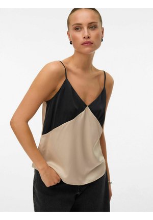 Kvinde iført sort og beige ærmeløs V-hals top med tynde stropper og sorte bukser, poserer med den ene hånd i lommen mod en hvid baggrund.