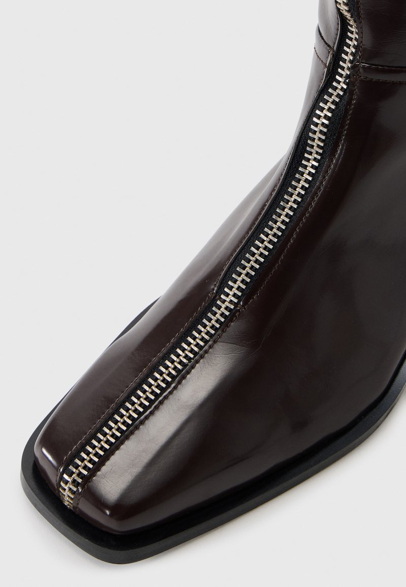 Botte cheville en cuir verni marron avec un bout carré, dotée d'une fermeture éclair visible et d'un accent noir contrastant sur la semelle.