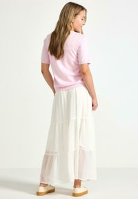Lys pink kortærmet sweater parret med en lang, lagdelt hvid nederdel. Nederdelen har gennemsigtige lag og et flagrende design, komplementeret af beige sko.