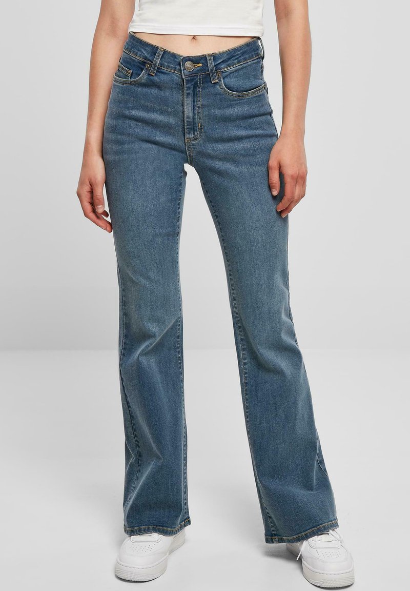 Flared jeans in light blue denim with a mid-rise waist, five-pocket design, and gold stitching, paired with white sneakers.