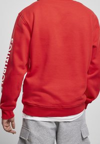 Uomo che indossa una felpa rossa con testo bianco sulle maniche e pantaloni cargo grigi, visto da dietro su uno sfondo neutro.