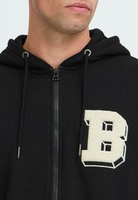 Uomo che indossa una felpa nera con zip e una grande lettera "B" bianca e strutturata sul lato sinistro del petto, con cordini dotati di punte in metallo.
