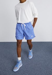 Osoba oblečená v bílé košili s dlouhým rukávem Nike, modrých Nike šortkách s tkaničkou, bílých ponožkách a modrošedých vysokých teniskách Nike, která jde uvnitř.