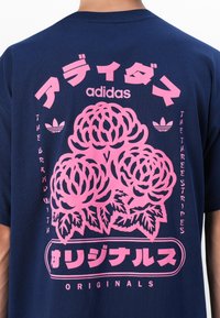 Tamsiai mėlynas medvilninis marškinėliai su didelėmis rožinėmis gėlių grafikomis ir "adidas" prekės ženklu ant nugaros, su tekstu tiek anglų, tiek japonų kalbomis.