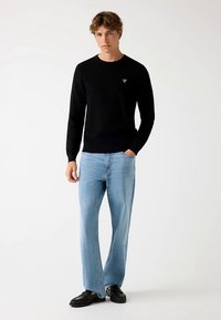 Sort strikket sweater med rund halsudskæring, lyseblå jeans med lige ben og sorte slip-on sko. Har et lille logo på brystet.