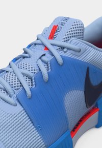 Αθλητικό παπούτσι σε ανοιχτό μπλε και λευκό με πλέγμα ύφασμα, μπλε επενδύσεις, μαύρο λογότυπο Nike και κόκκινες λεπτομέρειες στη γλώσσα και τη σόλα.