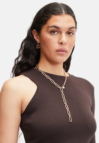 Guldtonet kæd necklaces med ovale led og en hængende accent, parret med matchende geometriske øreringe. Modellen har en mørkebrun top på.