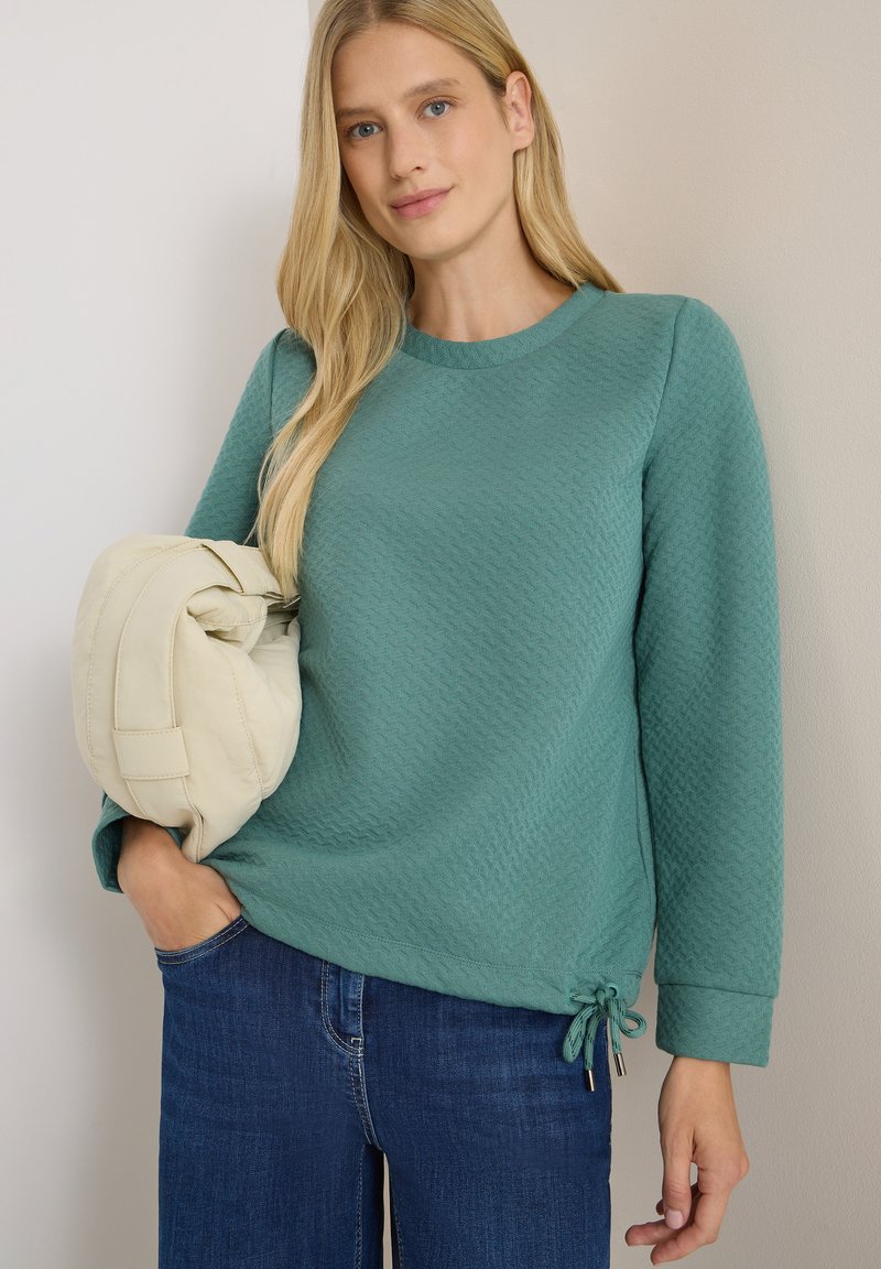 Kvinna iklädd en strukturerad grönblå långärmad tröja och mörkblå jeans, som håller en ljusbeige vadderad handväska under armen.