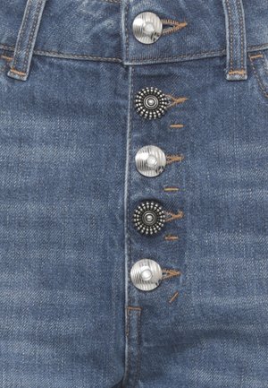Primo piano di jeans di denim blu con cinque bottoni decorativi in metallo, due neri con borchie e tre argento con scanalature, cuciti con filo marrone.